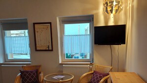Una sala de estar con dos sillones de mimbre, una mesita redonda, un mueble de madera y un televisor fijado a la pared.