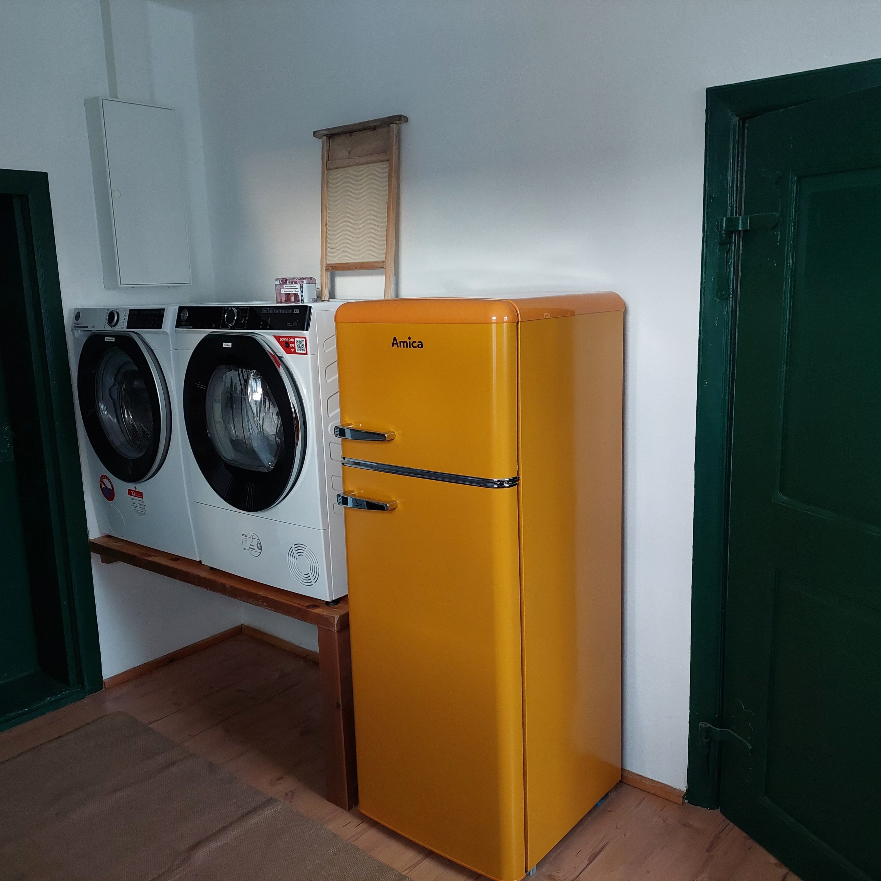 A compact room with a yellow Amica refrigerator, a washing machine, and a wooden shelf.