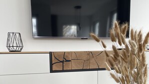 Ein modernes Wohnzimmer mit Flachbildfernseher an der Wand, einem Couchtisch aus Holz und einer Vase mit Pampasgras, sowie einem geometrischen Wandbild.