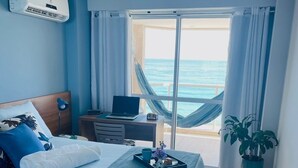 Una camera da letto con un letto, un portatile su un tavolino, una pianta in vaso e vista sull'oceano attraverso una grande finestra.