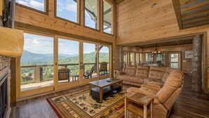 A spacious living room with a large wooden ceiling, a fireplace, a large sofa, and a view of the mountains through large windows. - Priceless Views, Indoor Pool Firepit Pool Table, Hot Tub, & Free Attractions! (Gatlinburg)