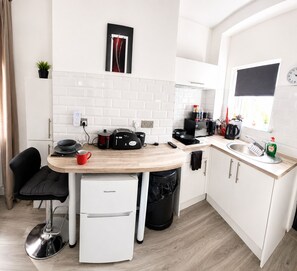 A compact kitchen with a white refrigerator, a small sink, a microwave, and a toaster. There is a black trash bin and a potted plant on the counter.
