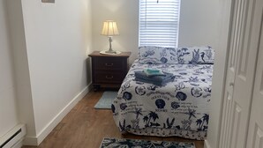 Un dormitorio con cama, mesita de noche, lámpara, ventilador de techo, ventana con persianas, alfombra estampada y puerta.