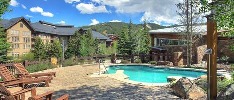 A pool area with lounge chairs, a stone patio, and a view of mountain buildings.
