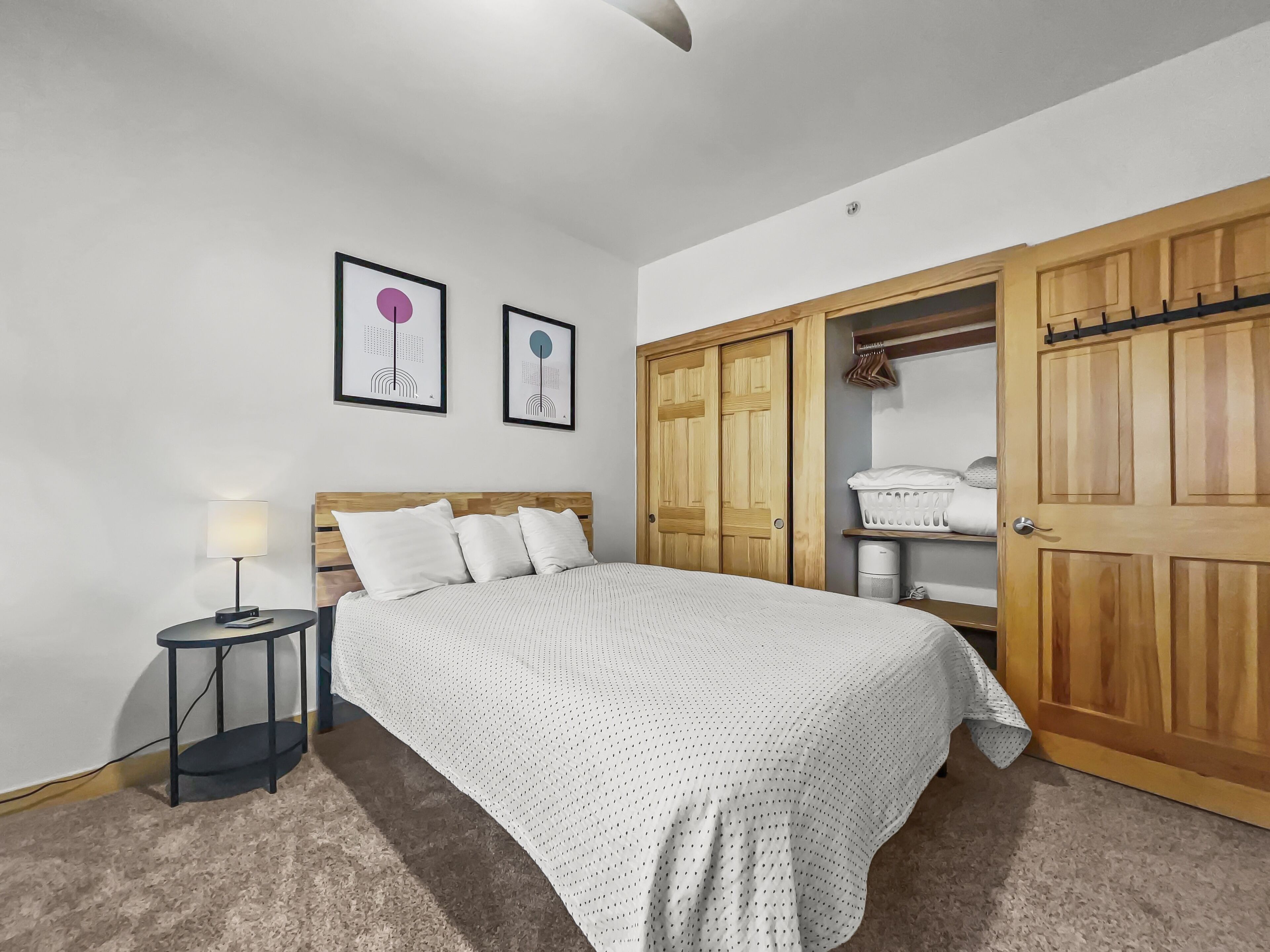 A bedroom with a bed, a nightstand, a lamp, a wardrobe, and two framed pictures on the wall.