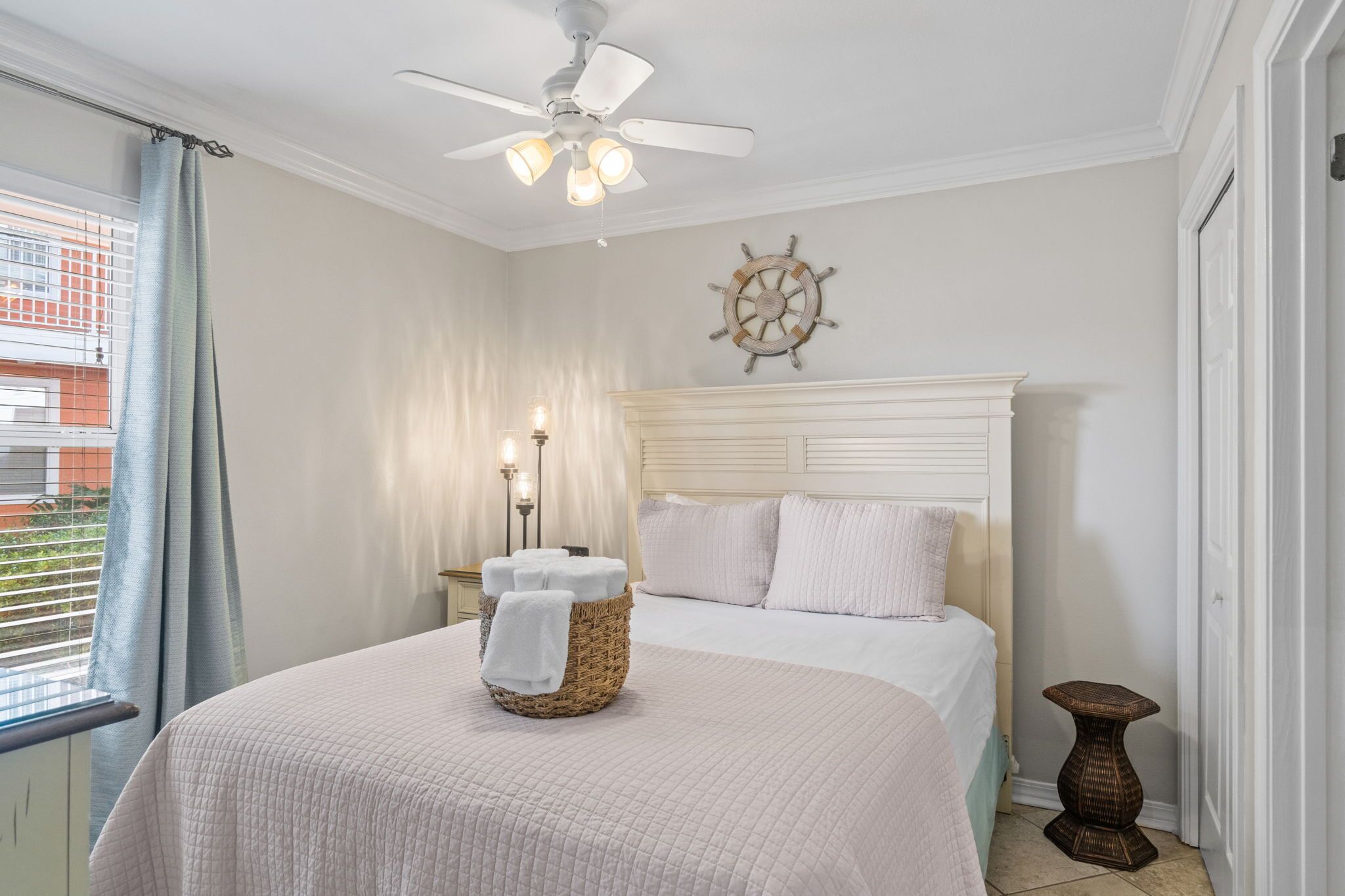 A bedroom with a bed, a ceiling fan, a ship's wheel decoration, and a wicker basket with towels.