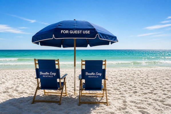 Two beach chairs with 'Breathe Easy Rentals' branding, a blue umbrella labeled 'FOR GUEST USE,' and a clear view of the ocean.