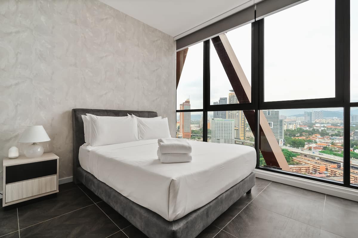 A modern hotel room with a large bed, a nightstand, and a view of the city through large windows.