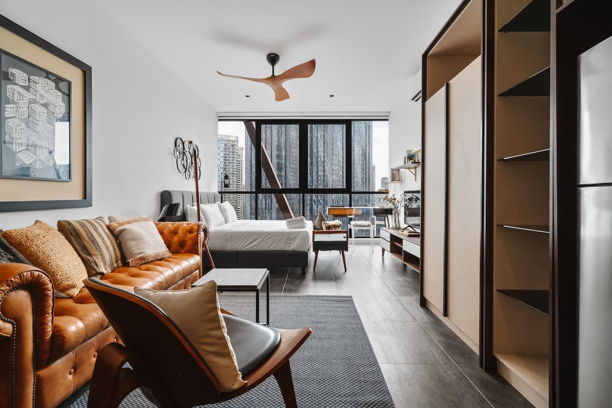 A modern hotel room with a large bed, a leather sofa, a dining area, and a view of the city skyline.