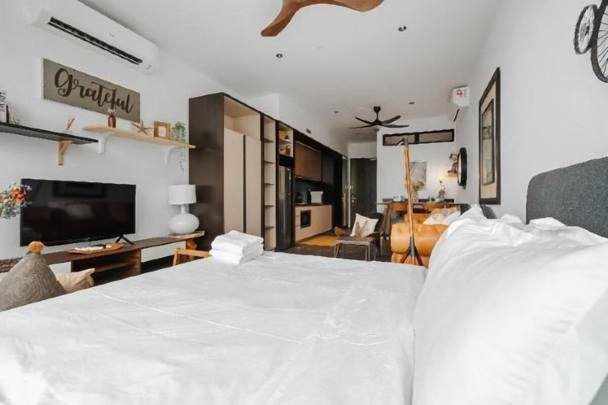A bedroom with a large bed, a television, a desk with a lamp, and a kitchenette in the background.