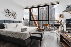 A modern bedroom with a large bed, a desk, and a view of tall buildings.