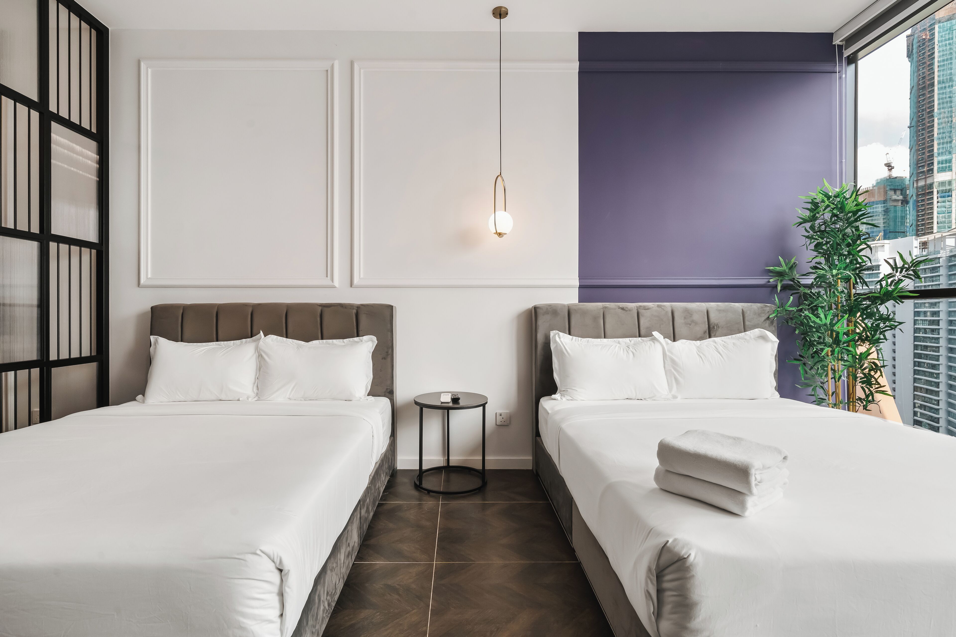 A hotel room with two beds, a small table, a wall-mounted light, and a view of the city.