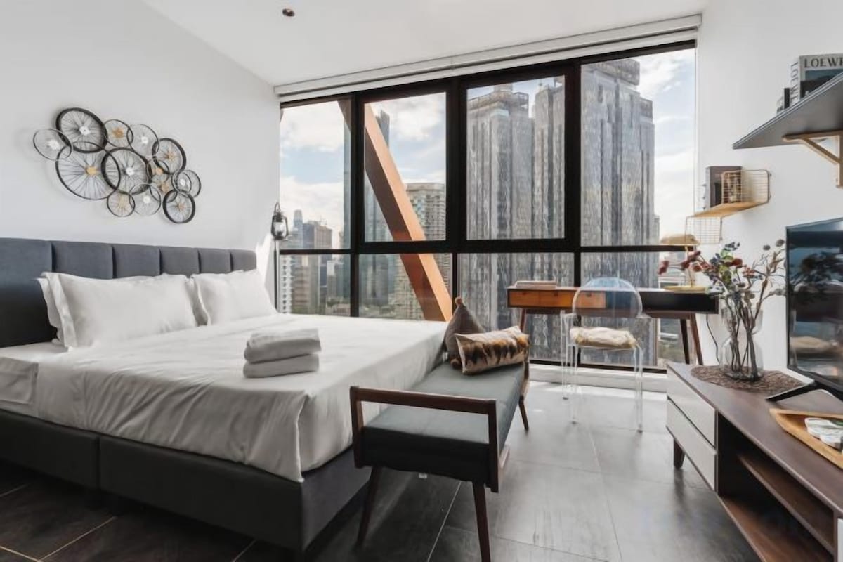A modern bedroom with a large bed, a desk, and a view of the cityscape.