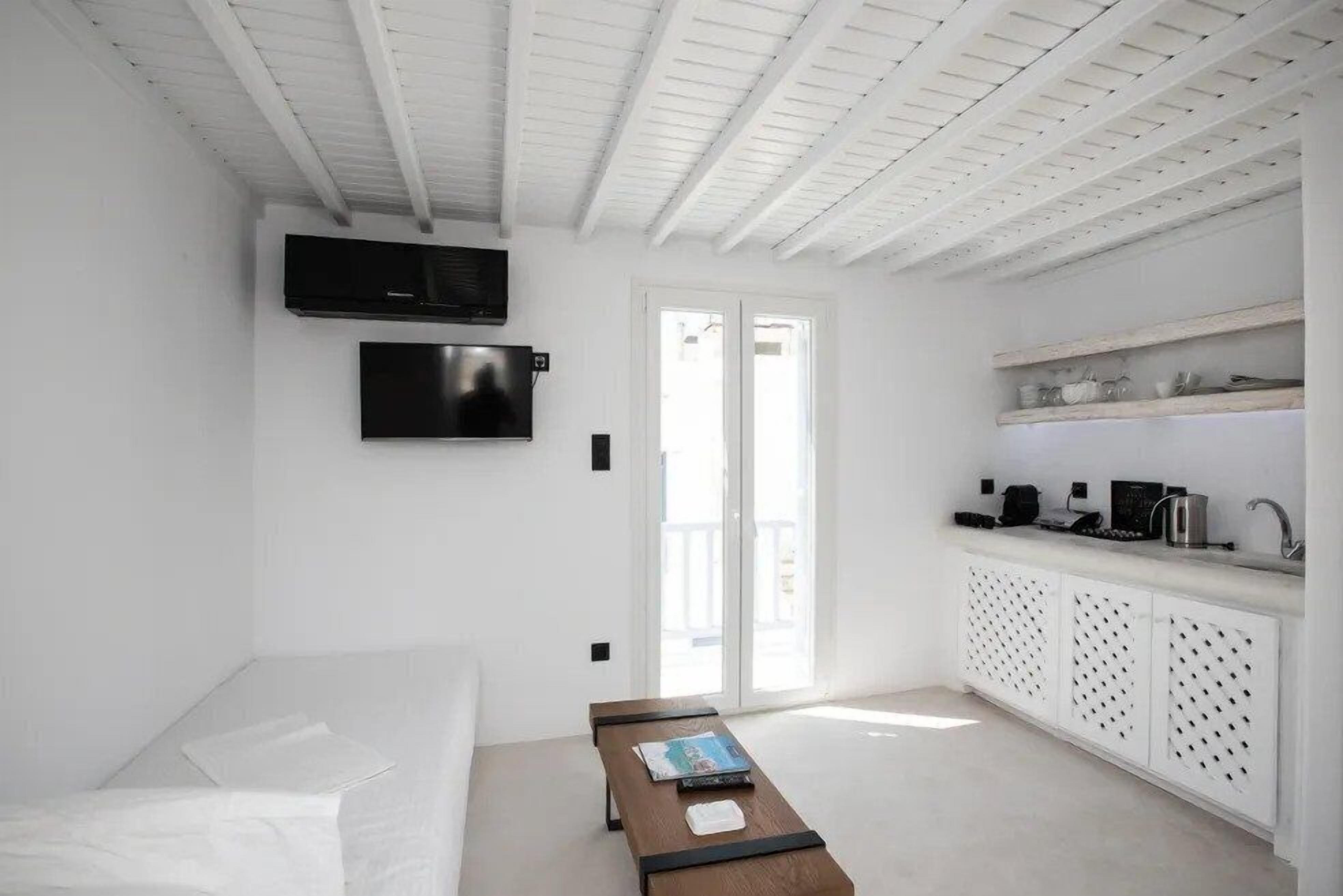 Une pièce au décor minimaliste, avec un canapé blanc, une table basse en bois, un téléviseur fixé au mur et un coin cuisine comprenant un évier et des étagères.
