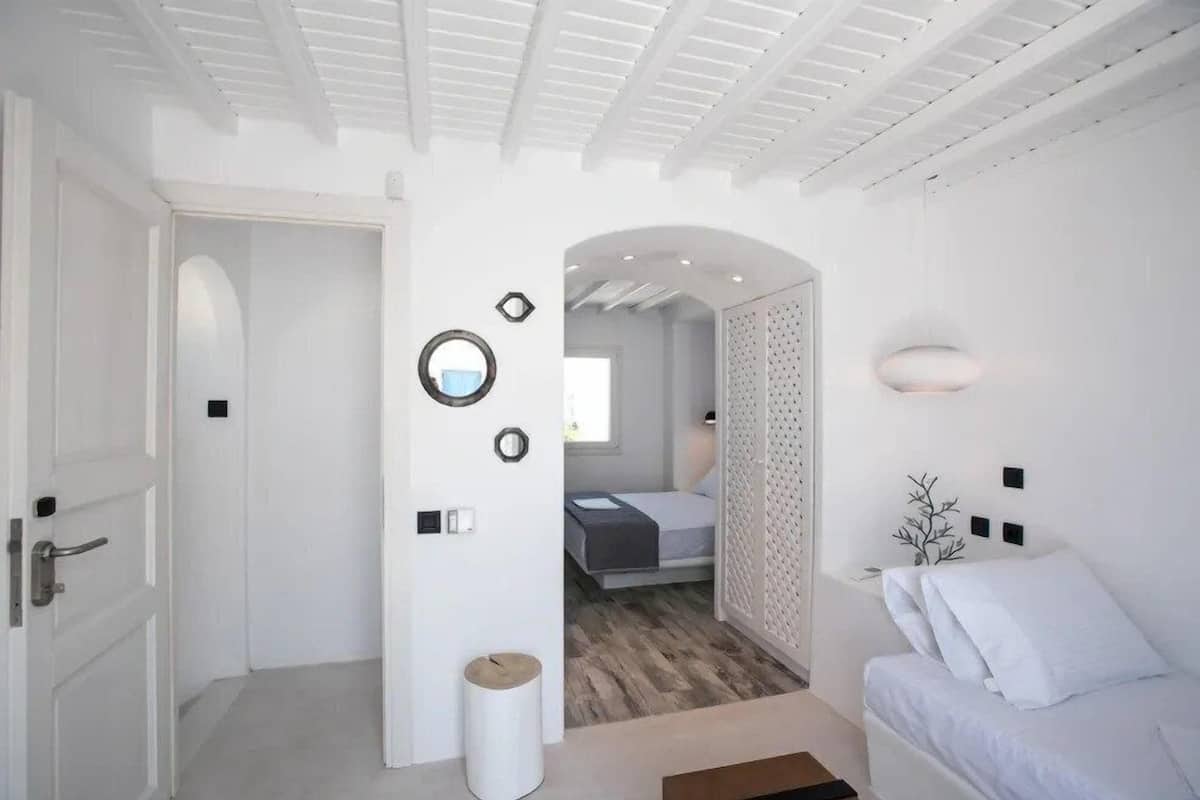 A modern, white-themed bedroom with a bed, a small bedside table, and a wall-mounted light fixture.