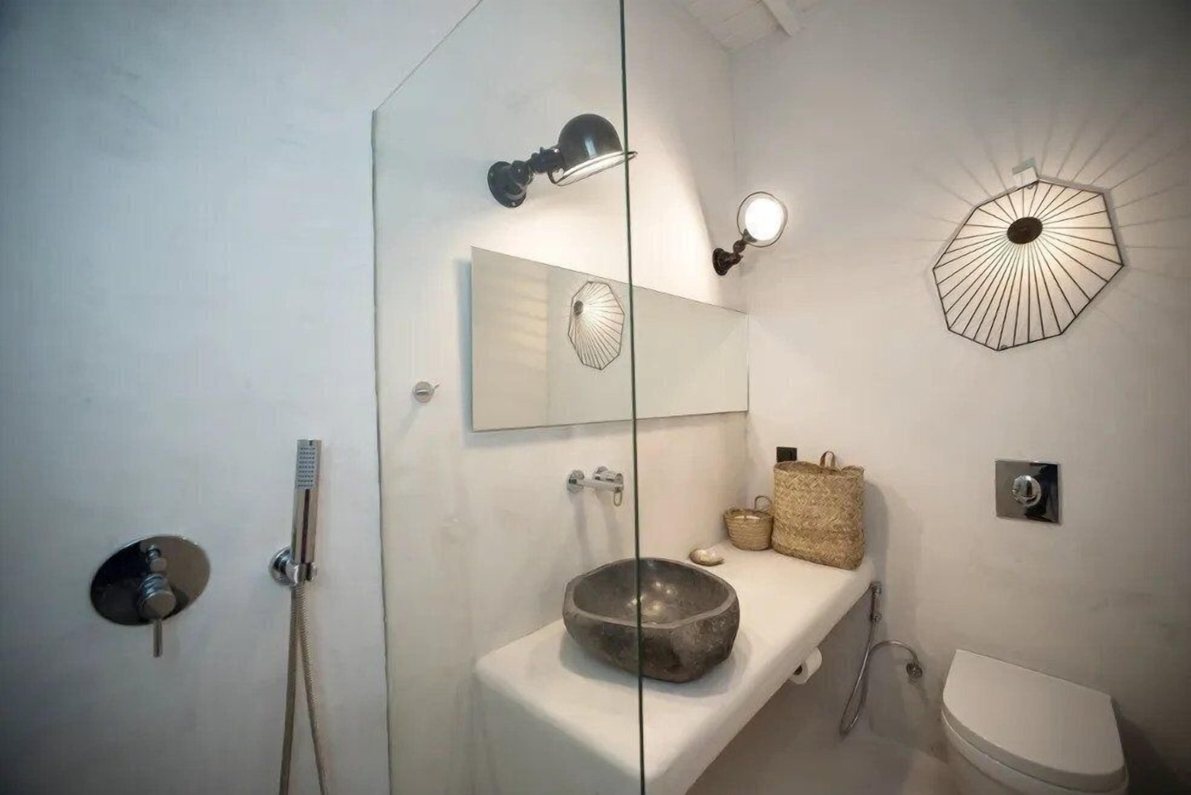 Un baño con cabina de ducha de vidrio, lavamanos de piedra, espejo, luz empotrada en la pared y retrete.