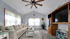 Un salón amplio con un ventilador de techo alto, un televisor de pantalla plana grande, una estantería de madera y un sofá cómodo.