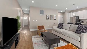 Una sala moderna con un televisor de pantalla plana, un sofá, una mesa de centro y un comedor con una cocina al fondo.