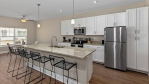 Una cocina moderna con armarios blancos, electrodomésticos de acero inoxidable, una isla de encimera de granito con taburetes altos e iluminación colgante.