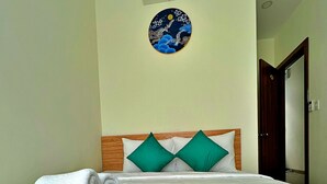 A neatly made bed with white linens and two green pillows, a wall-mounted air conditioner, and a circular artwork above the bed. - HANZ VeniceRiver Sunset Grand World (Phu Quoc)