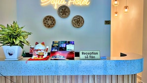 A hotel reception desk with a neon sign reading "Sofia Hotel," a plant, a cat figurine, and a sign indicating the reception area. - HANZ VeniceRiver Sunset Grand World (Phu Quoc)