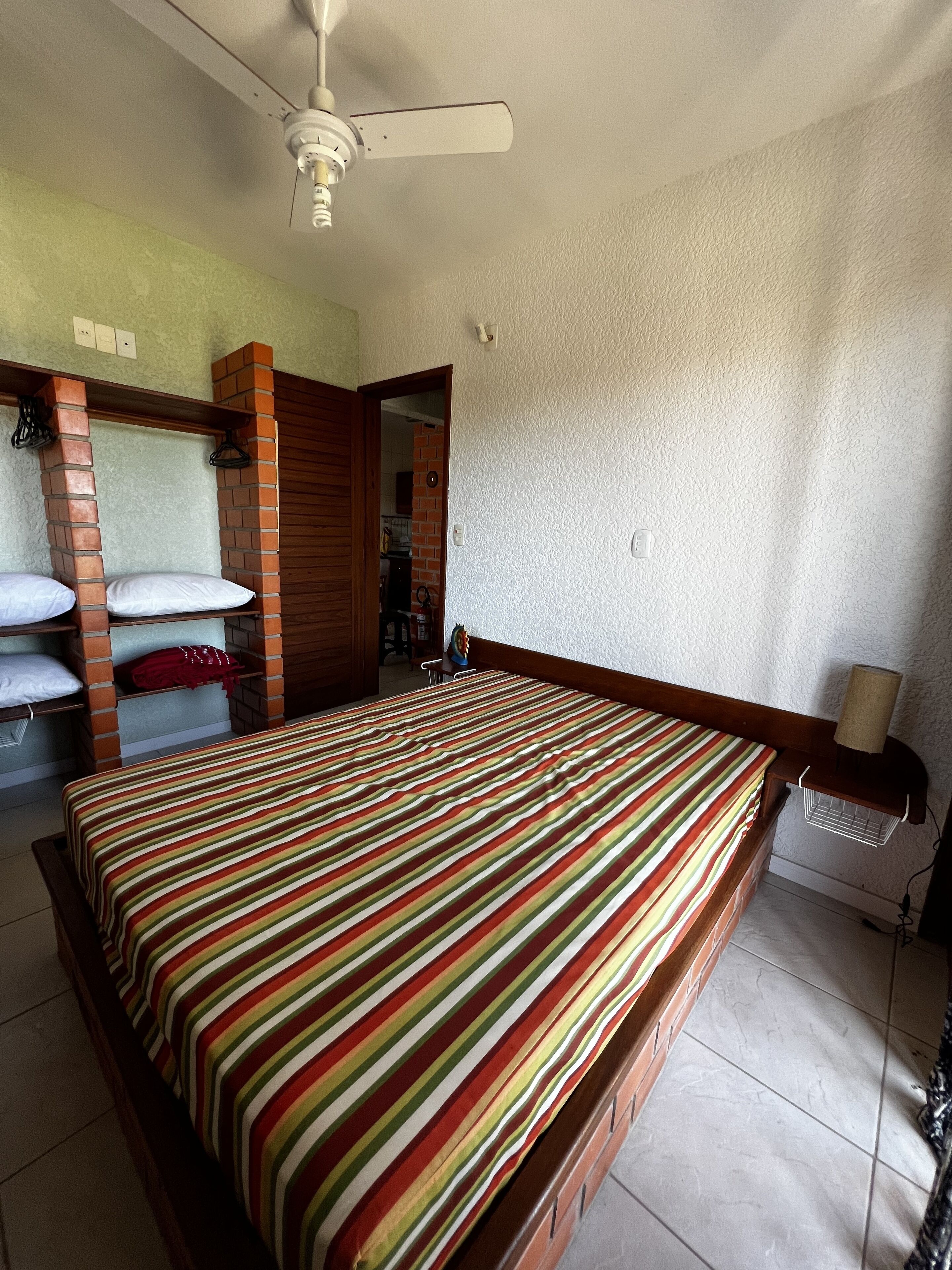 A bedroom with a bed, a ceiling fan, a brick wall, and a door leading to another room.