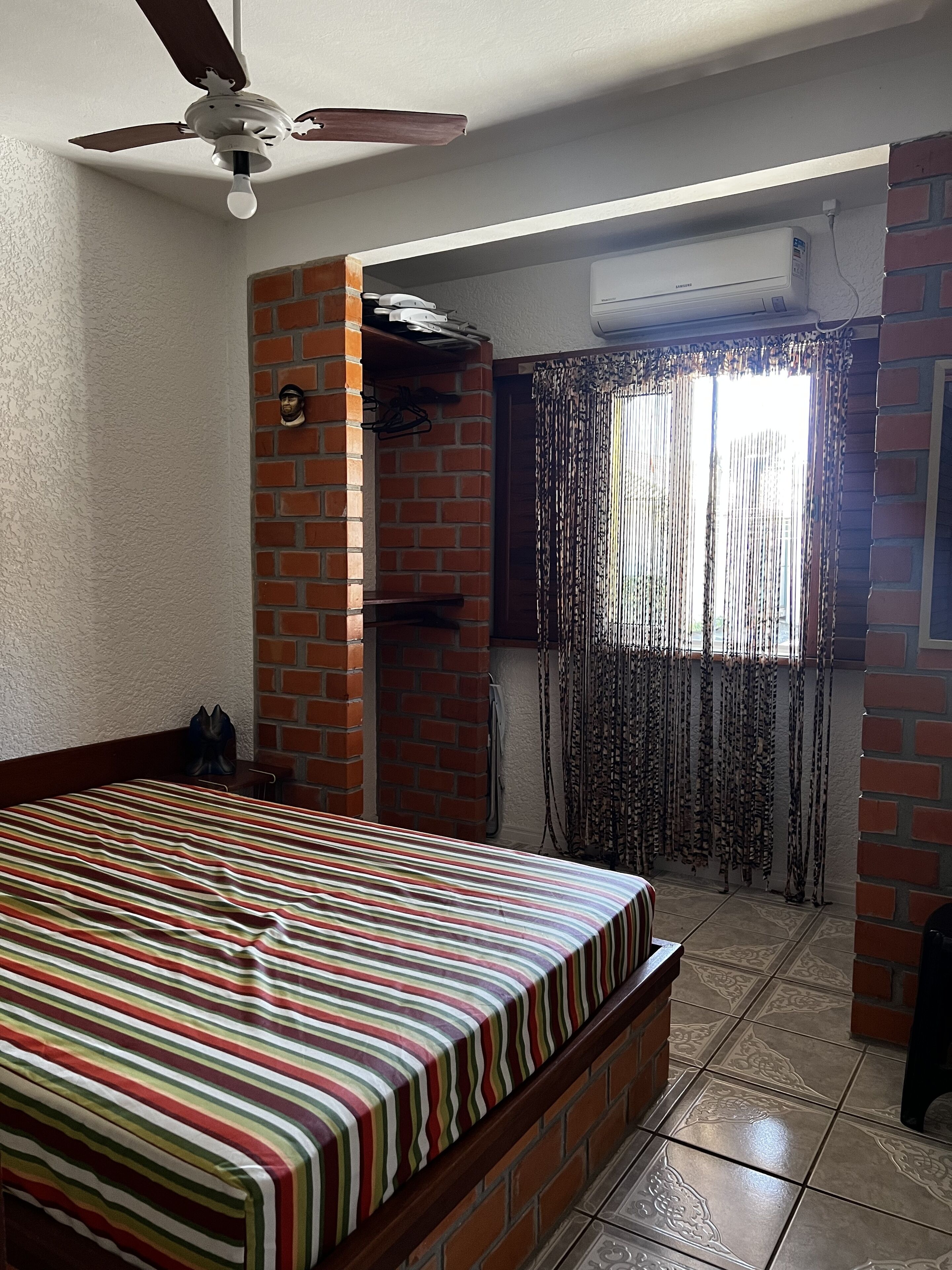 A bedroom with a bed, a ceiling fan, a brick wall, a window with curtains, and an air conditioning unit.
