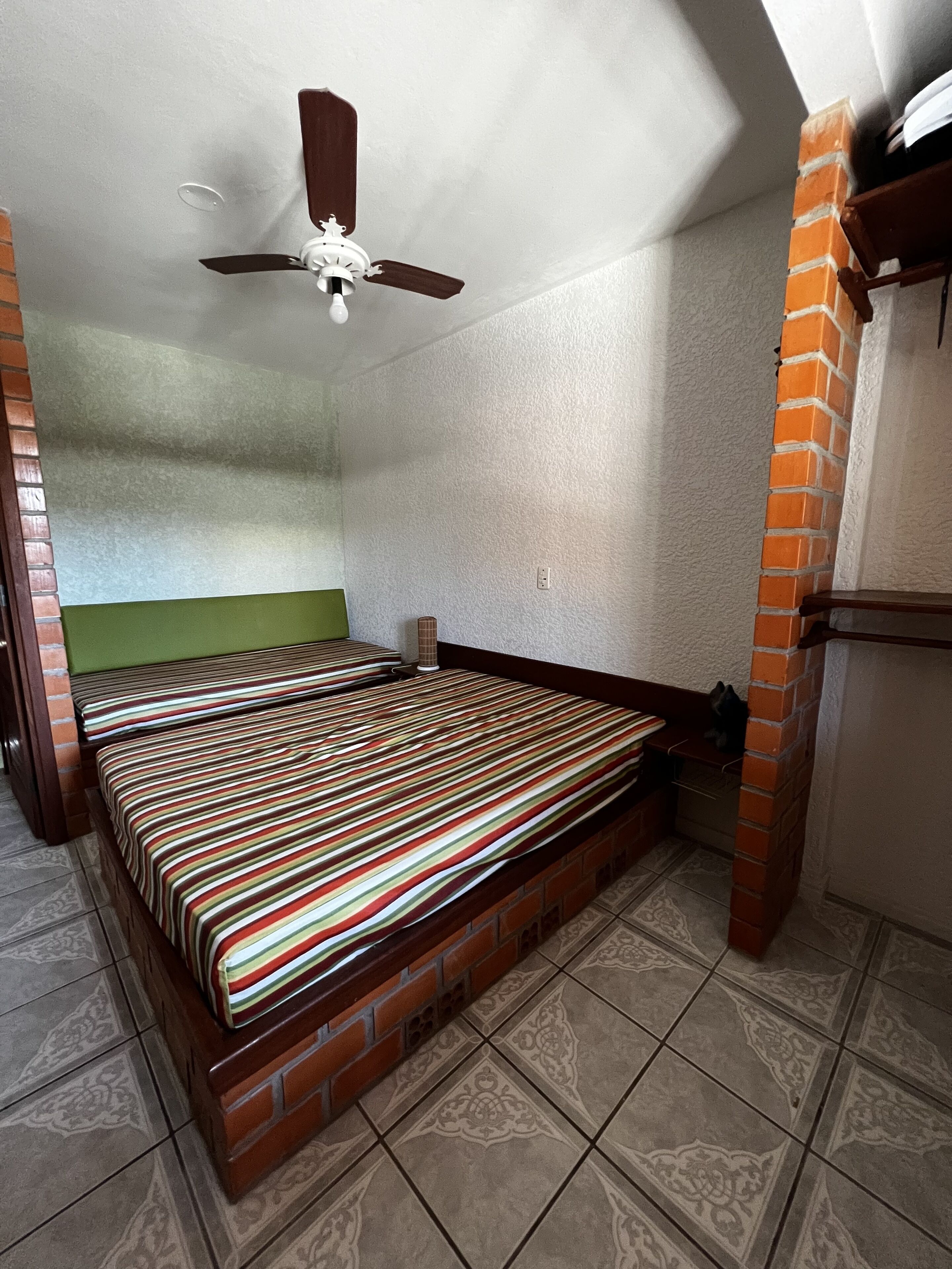 A room with a single bed, a ceiling fan, and a brick column.