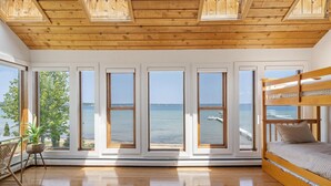 Une pièce avec un plafond en bois, de grandes fenêtres donnant sur la mer, un lit superposé et une plante.