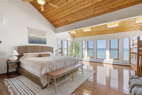 A spacious bedroom with a large bed, wooden ceiling, and large windows offering a view of the sea. - Loeb Lake House - sugar sand private beach - walking distance to Castle Farms (Charlevoix)