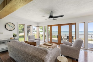 A spacious living room with a large window overlooking a deck and water. - Loeb Lake House - sugar sand private beach - walking distance to Castle Farms (Charlevoix)