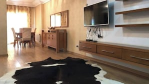 Een moderne woonkamer met houten vloer, een zwart-wit koeienhuidtapijt, een aan de muur bevestigde televisie en een houten kast met lades.
