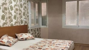 Een slaapkamer met een bed, twee ramen voorzien van jaloezieën, een behang met patroon en een houten vloer.