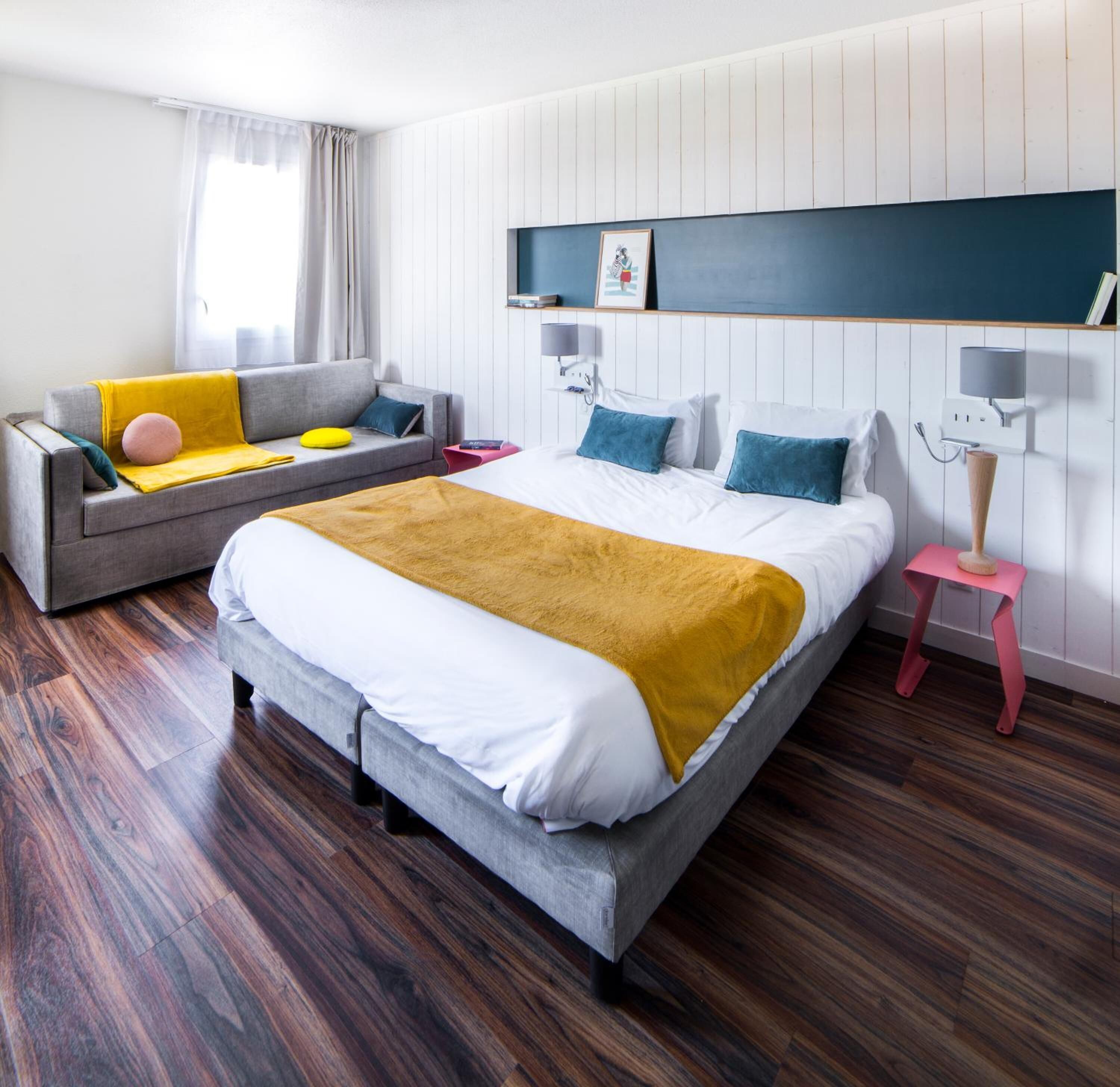 A modern hotel room with a large bed, a grey sofa with colorful cushions, a small pink side table, and a wooden floor.