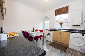 A compact kitchen with a washing machine, a dining area with red chairs, and a window with blinds.