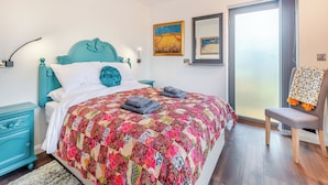 A bedroom with a teal headboard, a colorful quilt, a small bedside table, a chair with a patterned throw, and a view through a glass door.