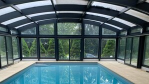 A covered swimming pool with a glass ceiling, surrounded by large windows offering views of trees and a car in the background. - Design villa with swimming pool and tennis court in a hectare of fully enclosed grounds (Le Pont-de-Beauvoisin)