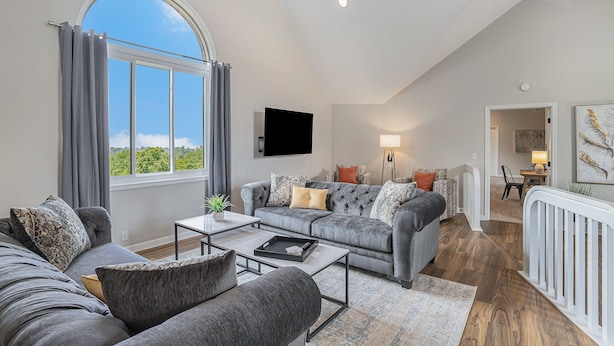 A spacious living room with a grey sofa set, a coffee table, and a large window with curtains. - Sunset Retreat Sleeps 30 | Hot Tub | Ping Pong | (Stevensville)
