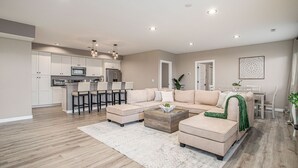 A modern living room with a sectional sofa, a coffee table, and a dining area with a kitchen in the background. - Sunset Retreat Sleeps 30 | Hot Tub | Ping Pong | (Stevensville)