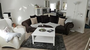 Ein Wohnzimmer mit einem braunen Sofa, einem weißen Couchtisch und einem weißen Sessel. Im Hintergrund ist eine Küche mit weißen Schränken und einer Mikrowelle zu sehen.