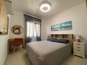 A bedroom with a bed, a desk, a chair, a nightstand, a lamp, a painting, and a ceiling light.