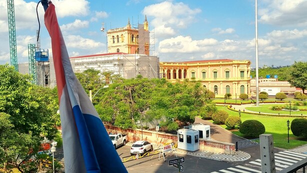 Vista de un paisaje urbano con un edificio destacado que tiene una torre de reloj, rodeado de follaje y un jardín bien cuidado.