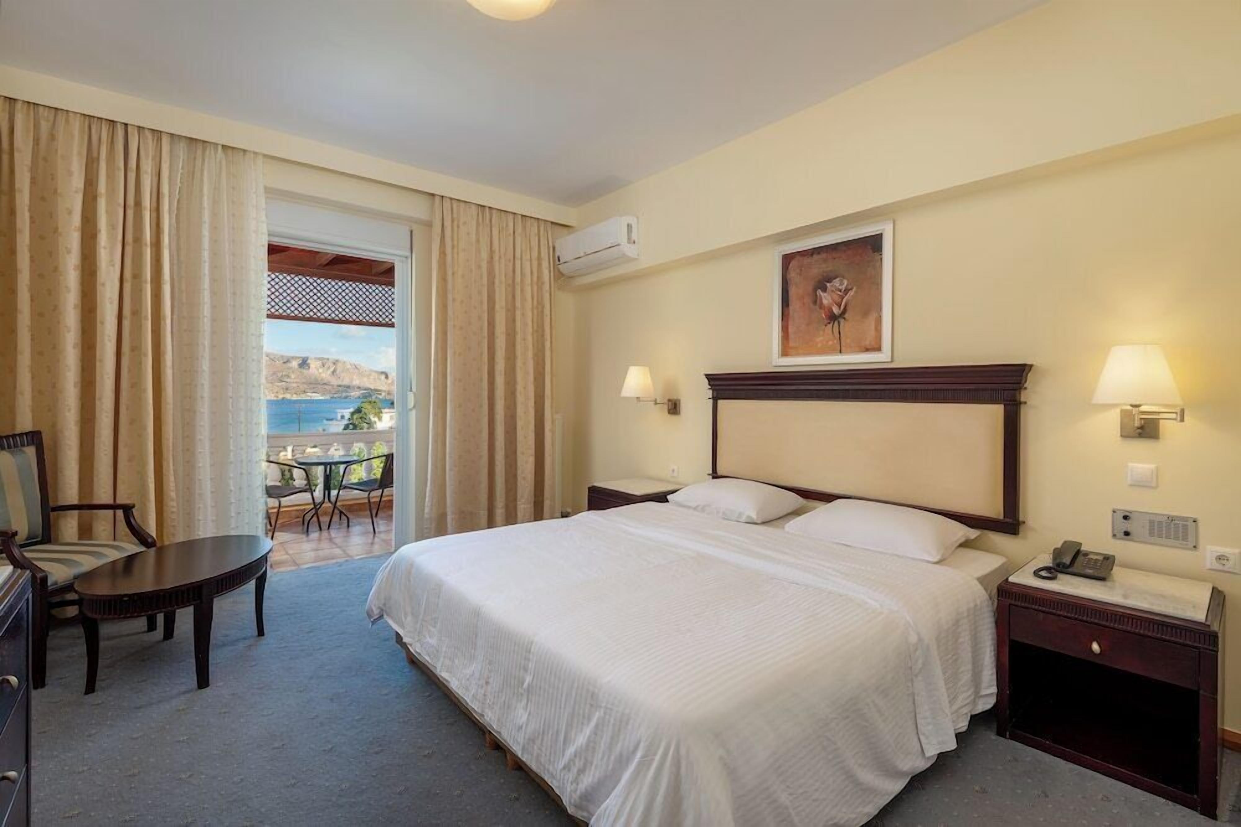 A hotel room with a large bed, a desk, a chair, a balcony with a view, and a painting on the wall.