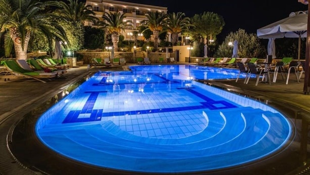 Una piscina con diseño geométrico, rodeada de sillones de descanso y palmeras, con un edificio al fondo.