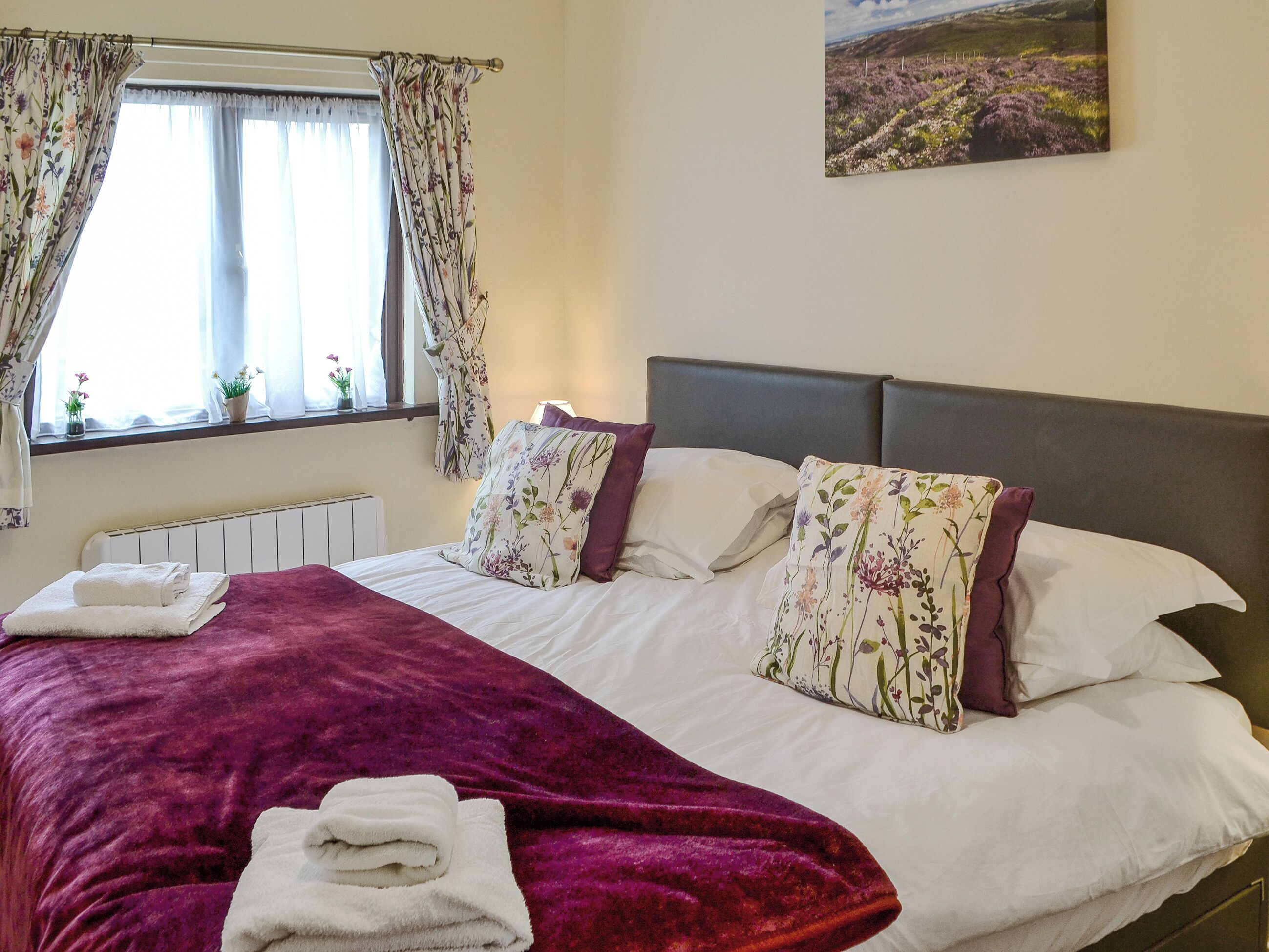 A neatly made bed with a purple blanket and white pillows, a window with floral curtains, and a painting of a landscape on the wall.