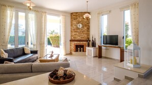 A living room with a stone fireplace, a sofa, a television, and a large window. - Cape Greco Villa Anastel 2 (Ayia Napa)