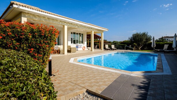 A swimming pool with a tiled deck, a covered patio area, and a view of a house in the background. - Cape Greco Villa Anastel 2 (Ayia Napa)