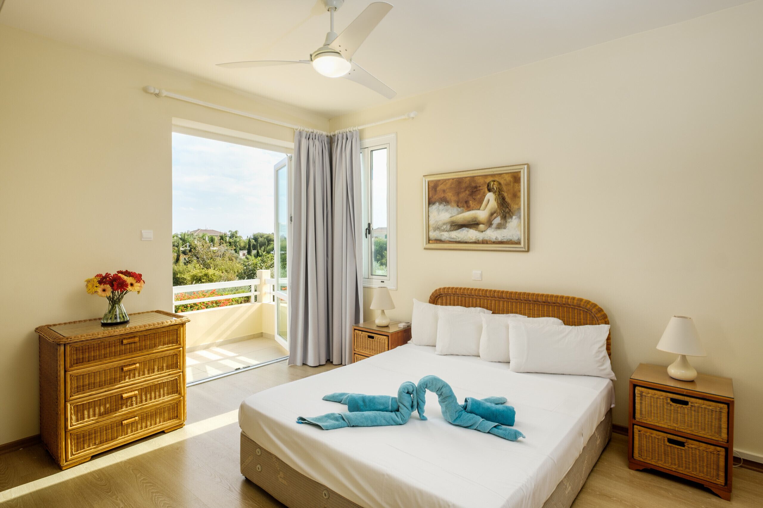 A hotel room with a bed, a ceiling fan, a painting, a nightstand, a lamp, a wicker side table, and a view of greenery outside.