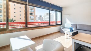 A modern hotel room with a large window, a sofa, a dining table, and a small white table.