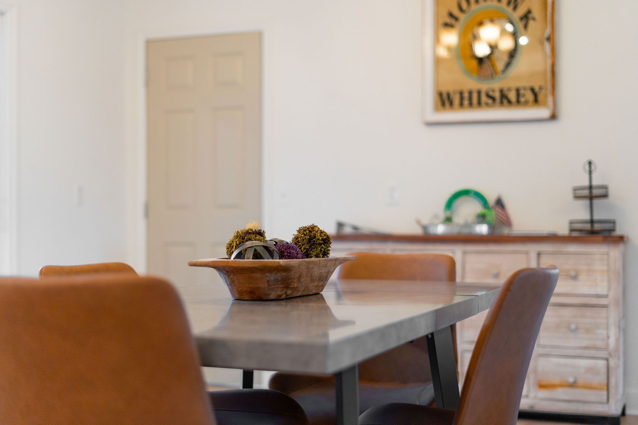Un coin repas avec une table dressée pour quatre personnes, un récipient contenant des objets décoratifs et une enseigne fixée au mur sur laquelle on peut lire : « MOWAN K WHISKEY ».